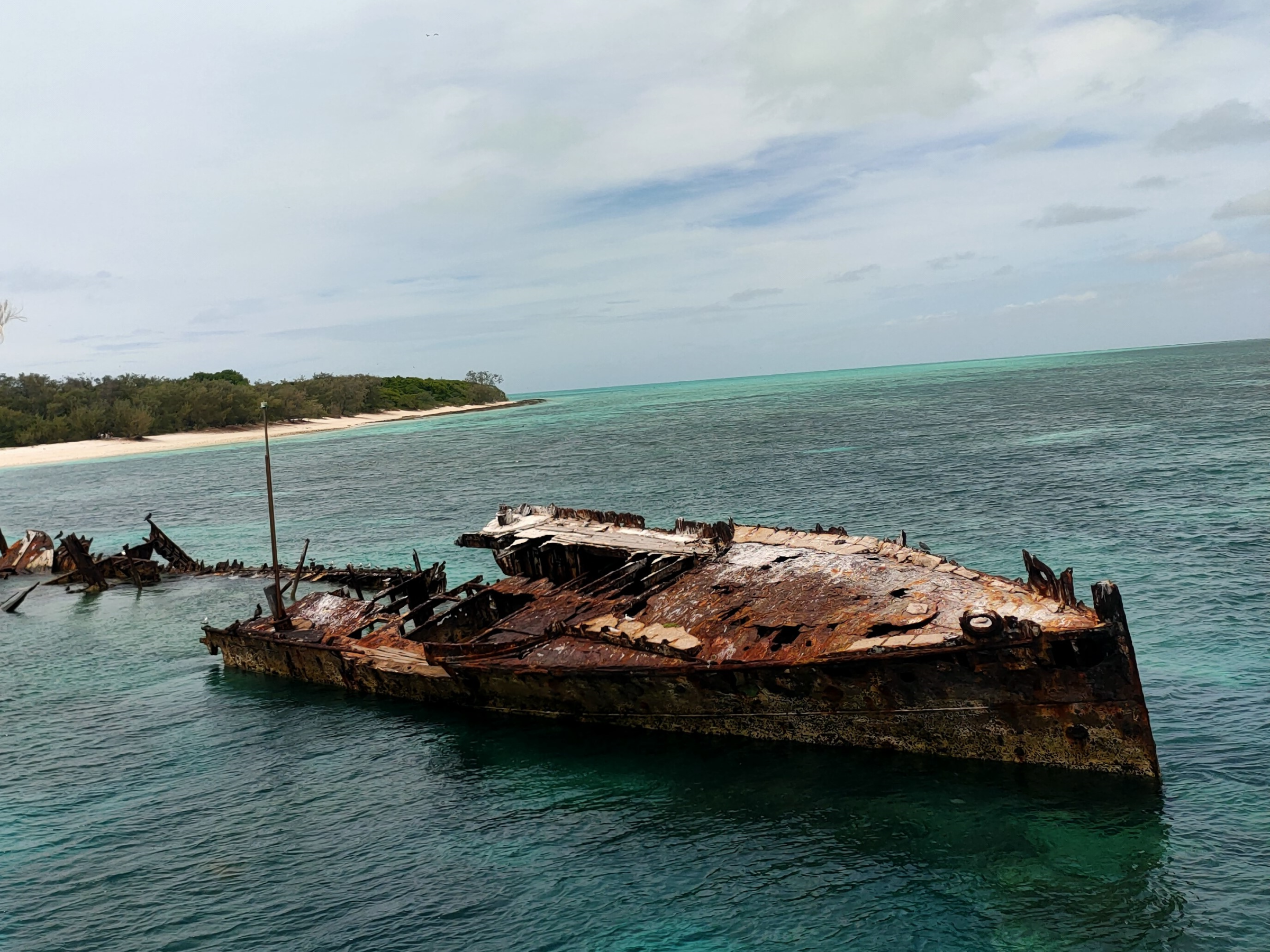 Rusty shipwreck in turquoise water near a sandy shoreline under a cloudy sky.